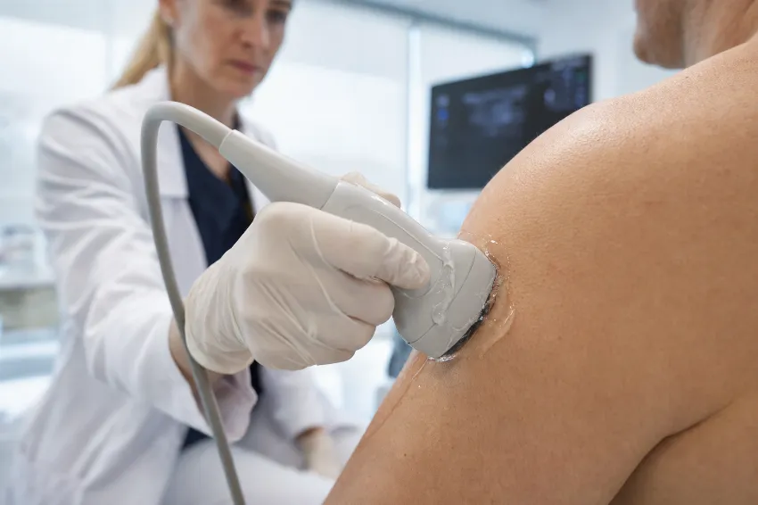 Detajl ultrazvočne sonde s prevodnim gelom, prislonjene na ramo pacienta med diagnostiko mehkih tkiv.
